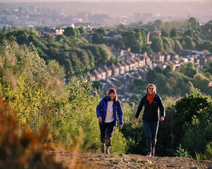 Two ramblers are walking up a hill. Down in the valley below them you can see rows of terrace houses and on the horizon you can see Sheffield city centre.