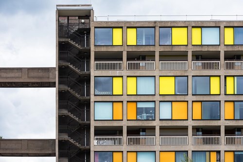 A section of a modernist apartment building with a concrete exterior and a grid of square windows in bright yellow, orange, and light blue panels. The left side features an open stairwell with metal railings and a connecting walkway. The overall design is geometric and colourful, contrasting with the cloudy sky in the background.