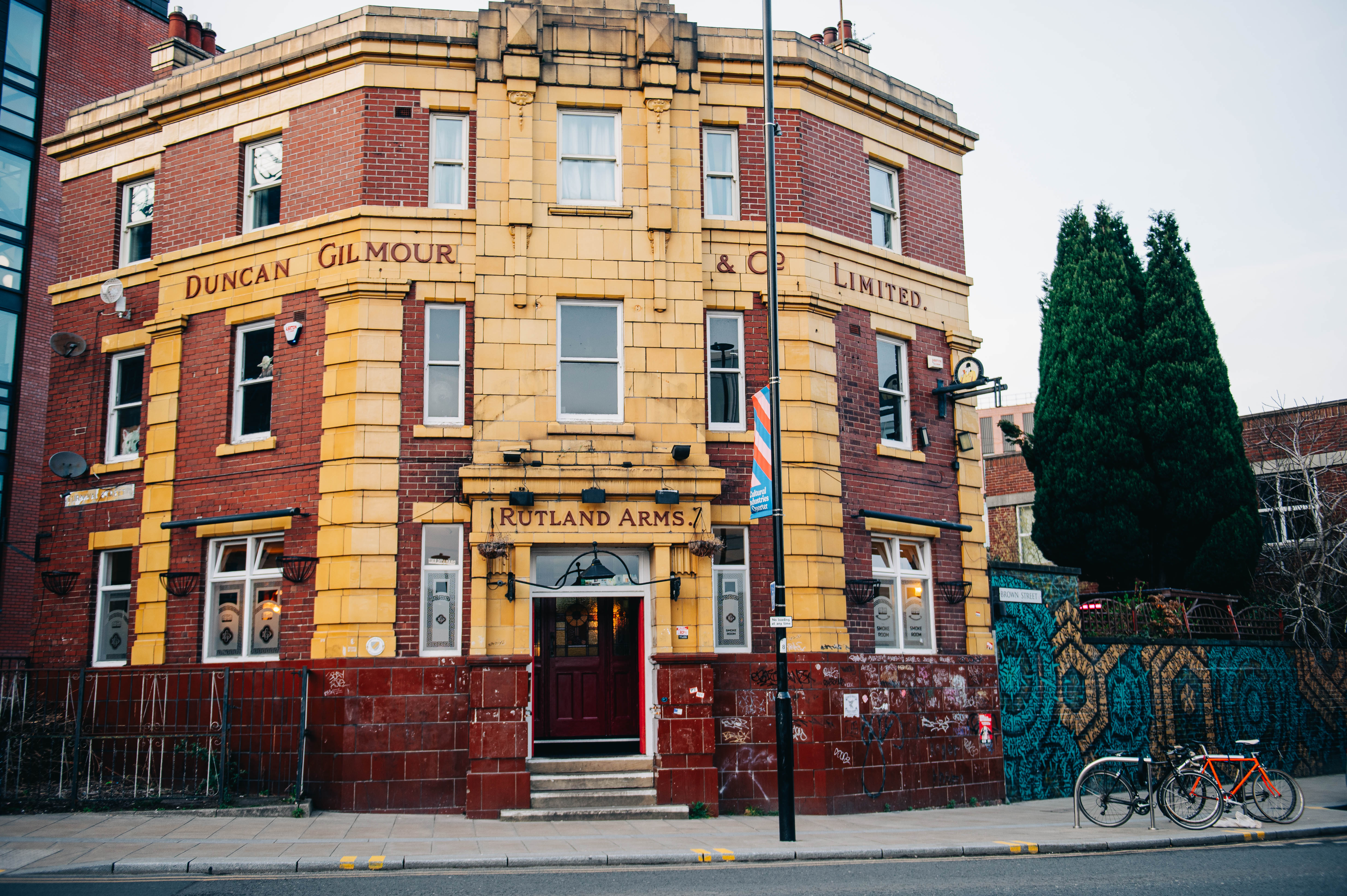 Rutland Arms exterior. The building is red and mustard brick, with grey sky in the background and two bicycles and street art to the right of the building.
