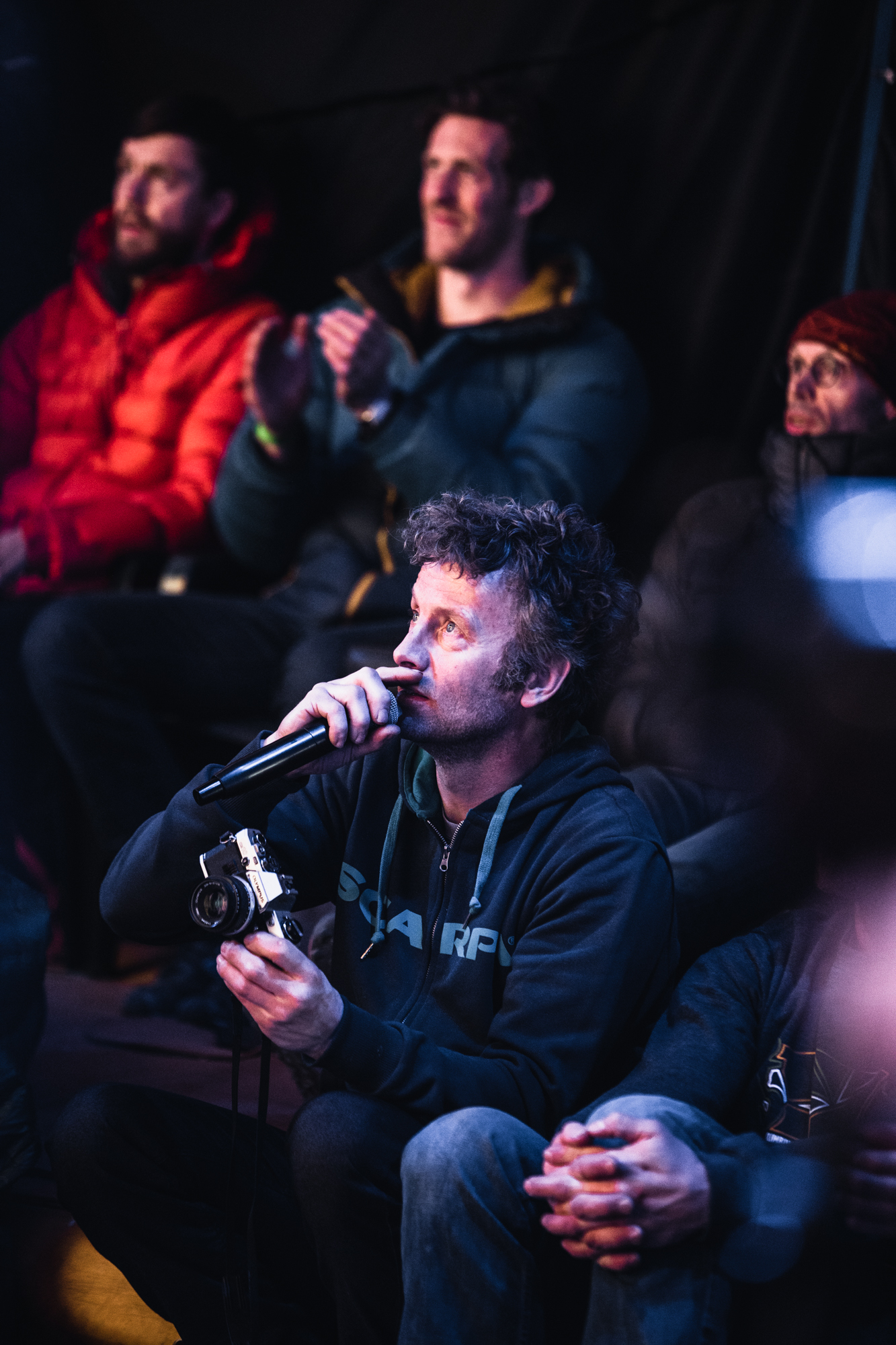 A man is sat in a group holding a microphone and a camera.