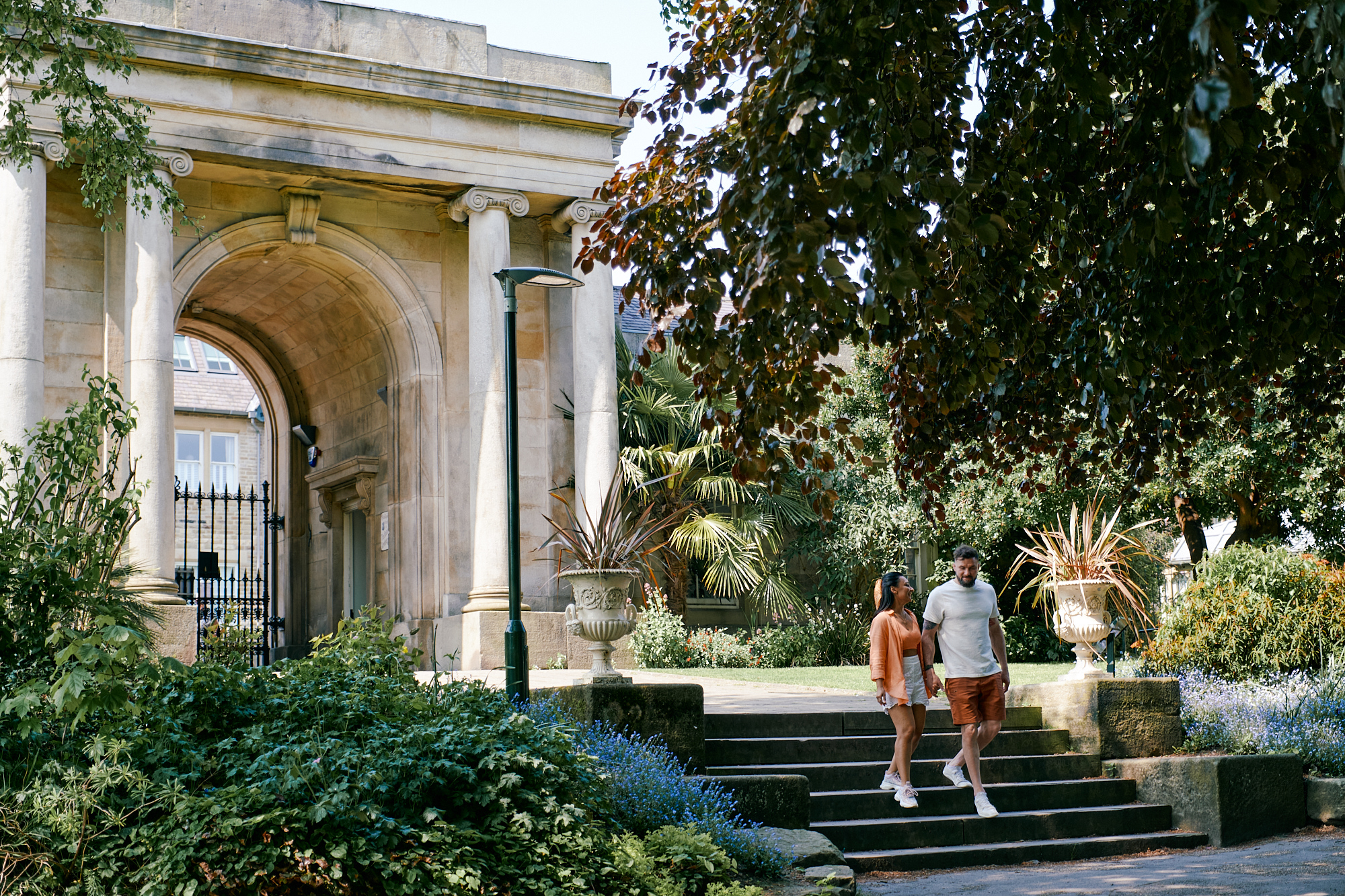 Couple walk down steps with the grand archway at the entrance to Sheffield Botanical Gardens in the background