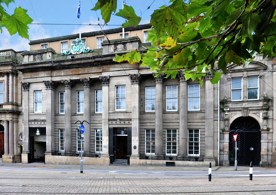 The exterior of Cutlers' Hall. A historic stone building with neoclassical architecture featuring tall columns, large rectangular windows, and ornate detailing above the entrance. The facade includes decorative carvings and a crest at the top center. Tram tracks run along the street in front, and green leaves from a nearby tree frame the upper part of the image.