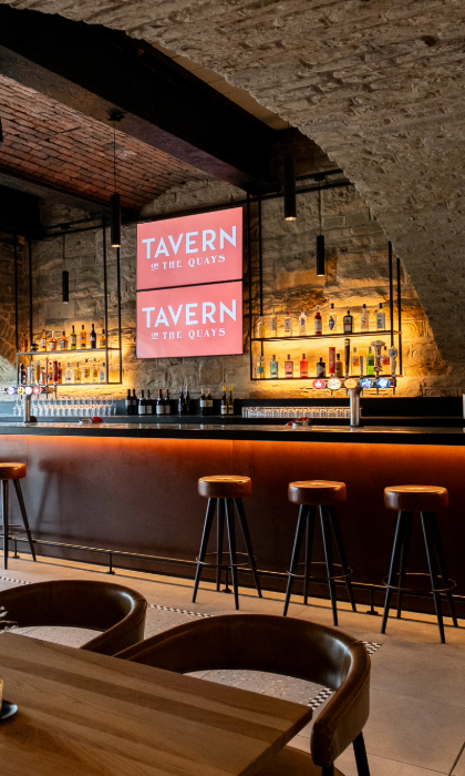The bar at Tavern on The Quays, with the vaulted stone ceiling plus bar stools lined up along the bar and tables and chairs in the foreground.
