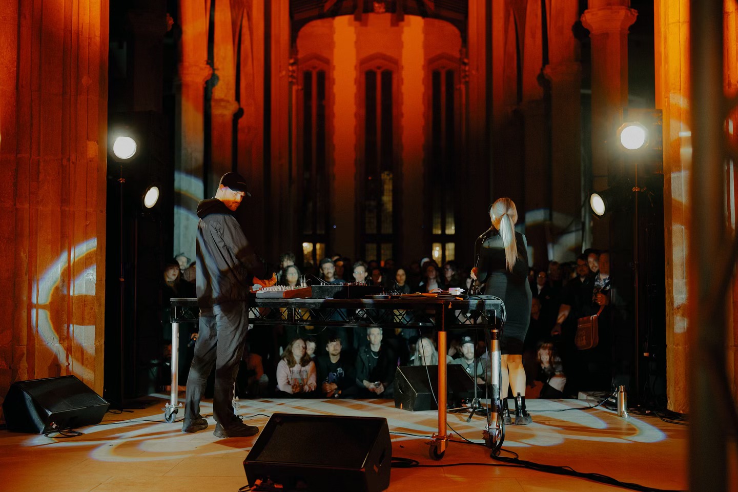 Two performers stand behind a DJ table in a grand hall with tall columns and arched windows, illuminated by warm orange lighting and spotlights. A seated audience watches in the background.