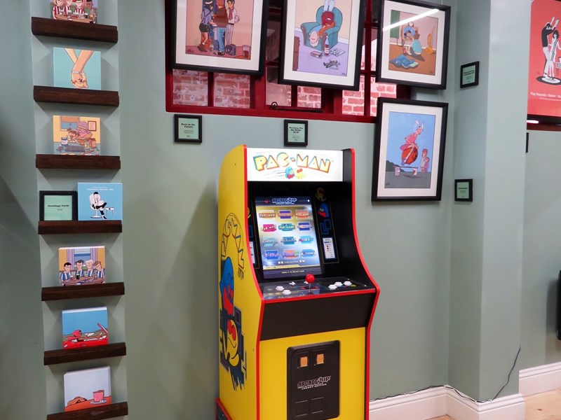 A Pac-Man arcade machine in the Pete Mckee Gallery, surrounded by prints on the wall.