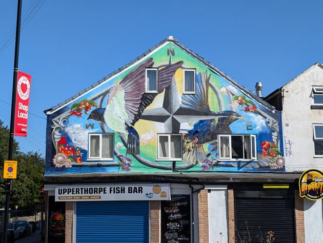 Large mural on the upper facade of a building featuring two birds in flight around a compass rose with letters N, E, S, W. The artwork includes colorful flowers and a bright blue sky background. Below the mural is a shop sign reading ‘Upperthorpe Fish Bar’ with closed blue shutters.