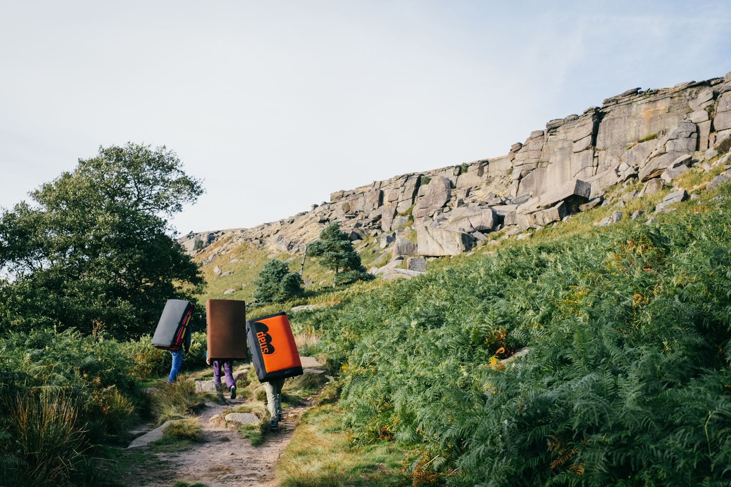 Three people are walking along a path that leads to a huge gritstone rock face. They are all carrying climbing crashmats on their backs.