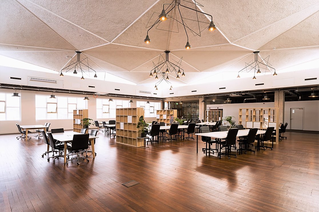 A spacious, modern office with an open floor plan, high ceilings featuring geometric patterns, and hanging light fixtures. Tables and chairs are arranged in clusters for collaborative work. The polished wooden floor and large windows provide a bright, welcoming atmosphere. Bookshelves and potted plants are placed throughout, enhancing both aesthetics and accessibility.