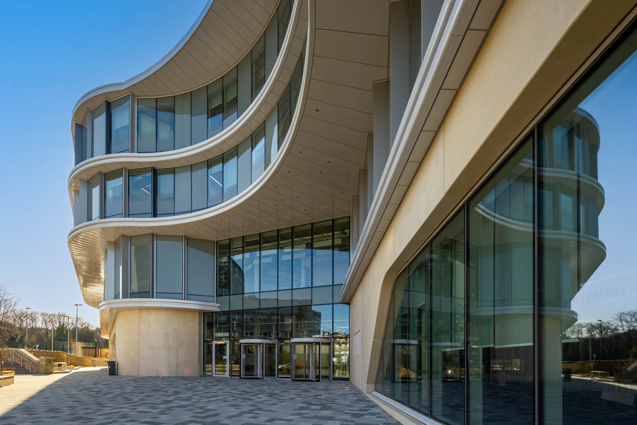 Modern building with a striking curved glass façade and multiple levels featuring sweeping architectural lines. The entrance has large glass doors beneath an overhang, and the exterior includes reflective windows and light-coloured panels. The paved area in front of the building leads to an open outdoor space under a clear blue sky.
