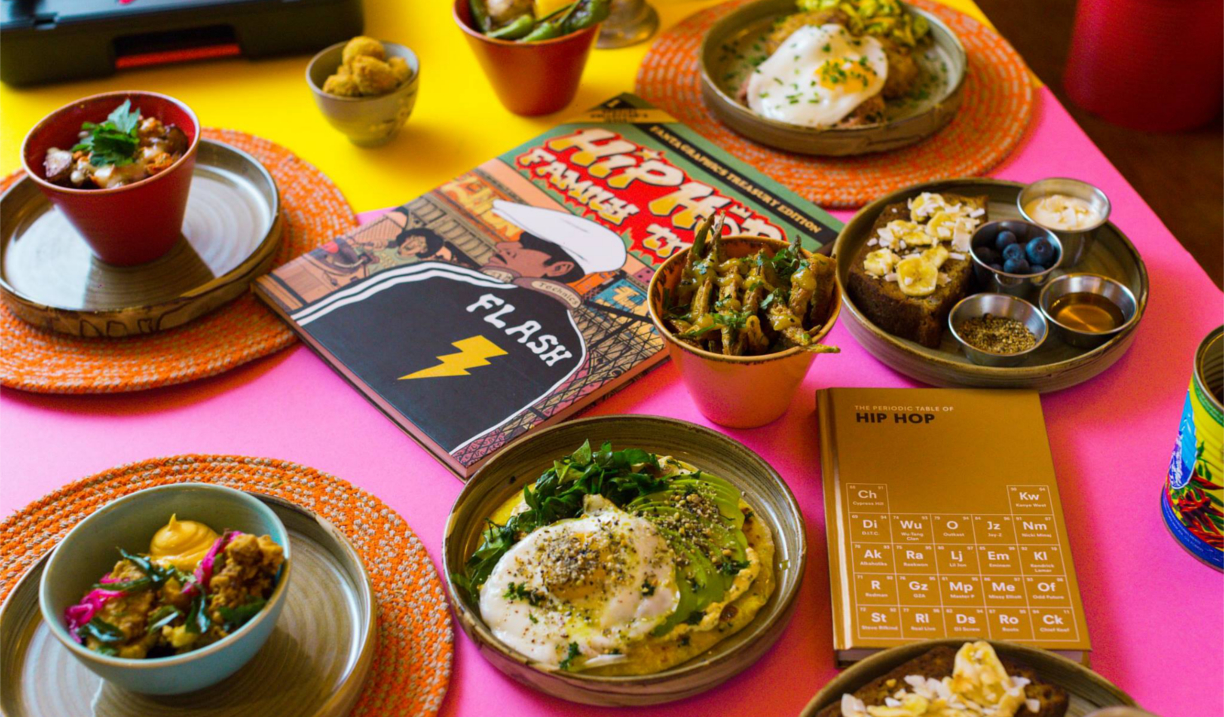 A colorful table setting featuring several plates and bowls of assorted dishes, including eggs on greens, fried vegetables, and small sides with sauces and blueberries. Two illustrated hip-hop themed books are placed among the food, and the table is covered with bright pink and yellow surfaces accented by orange placemats.