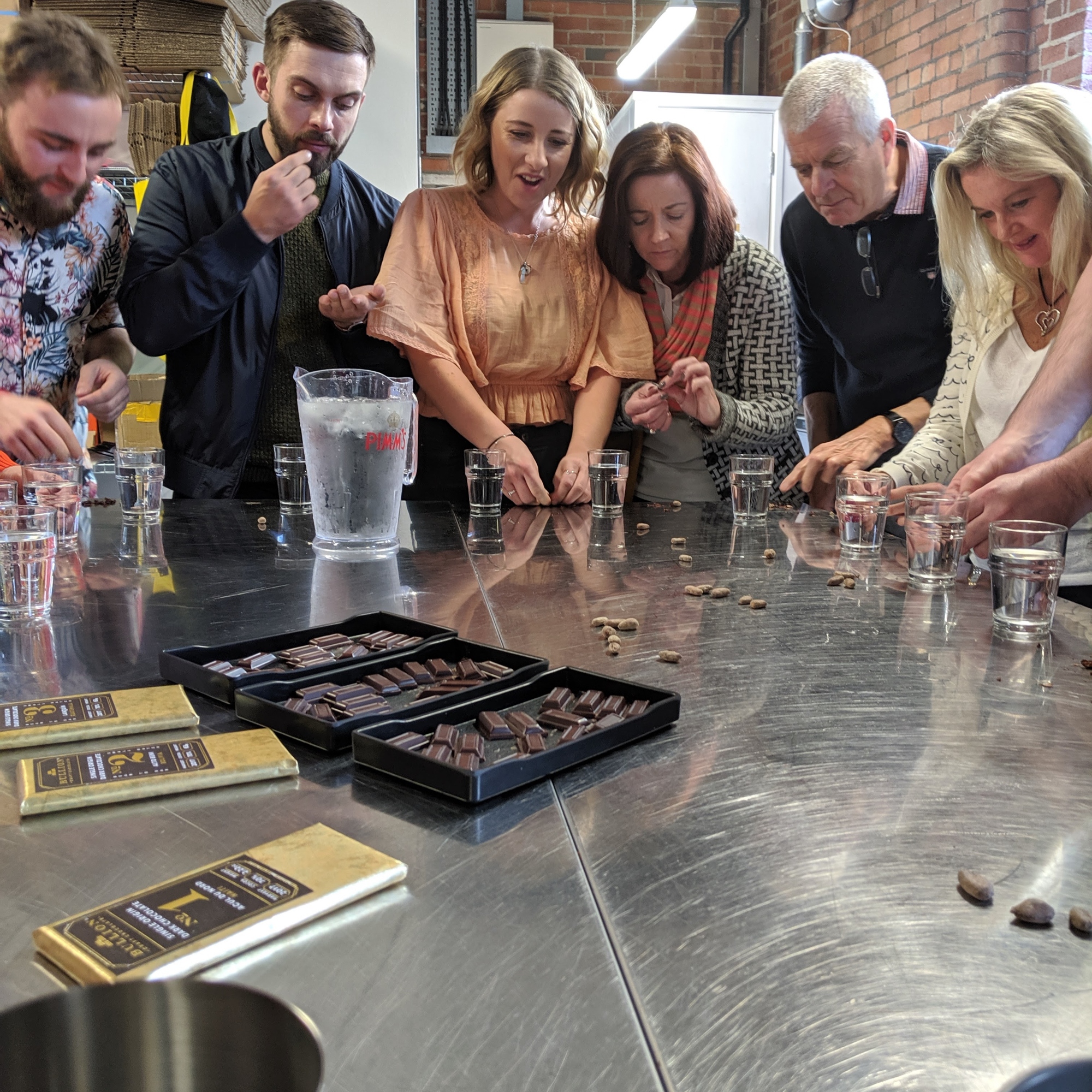 Group of people standing around a stainless steel table during a chocolate tasting or workshop. The table has trays of assorted chocolate pieces, several chocolate bars, and clear glasses. A large water jug is also visible. The setting appears to be an industrial-style kitchen with brick walls and shelves in the background.