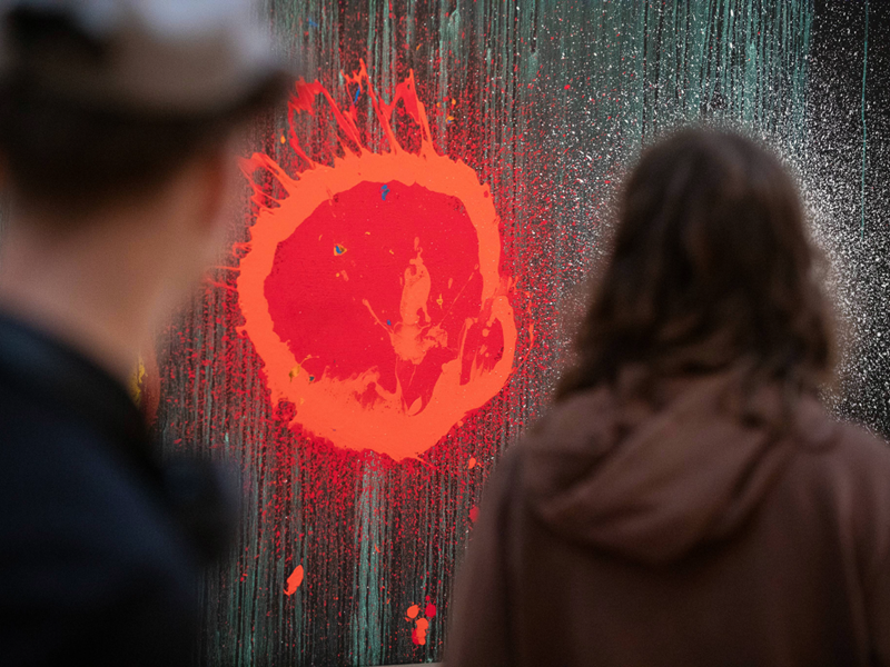 Two people stand observing a piece of abstract art. The artwork features a vivid red splatter resembling an explosion or sunburst, set against a dark, textured background with streaks of green and white. The red element is the focal point, creating a striking contrast and dynamic visual impact.