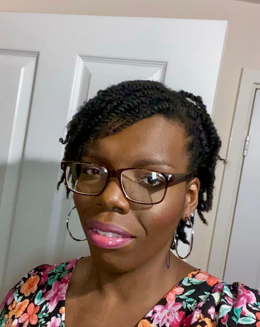 Close-up of a person wearing a floral-patterned top with pink, green, and orange flowers against a dark background. The image focuses on the upper body and hairstyle.