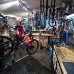 A photograph of the interior of The Bike Garage with the walls covered in tools and spare parts. There are two men working on bikes.