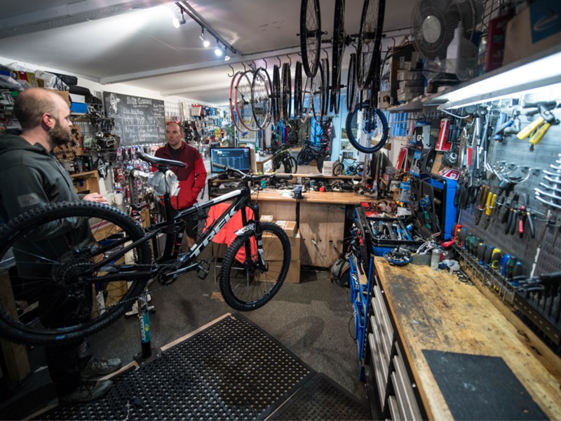 A photograph of the interior of The Bike Garage with the walls covered in tools and spare parts. There are two men working on bikes.