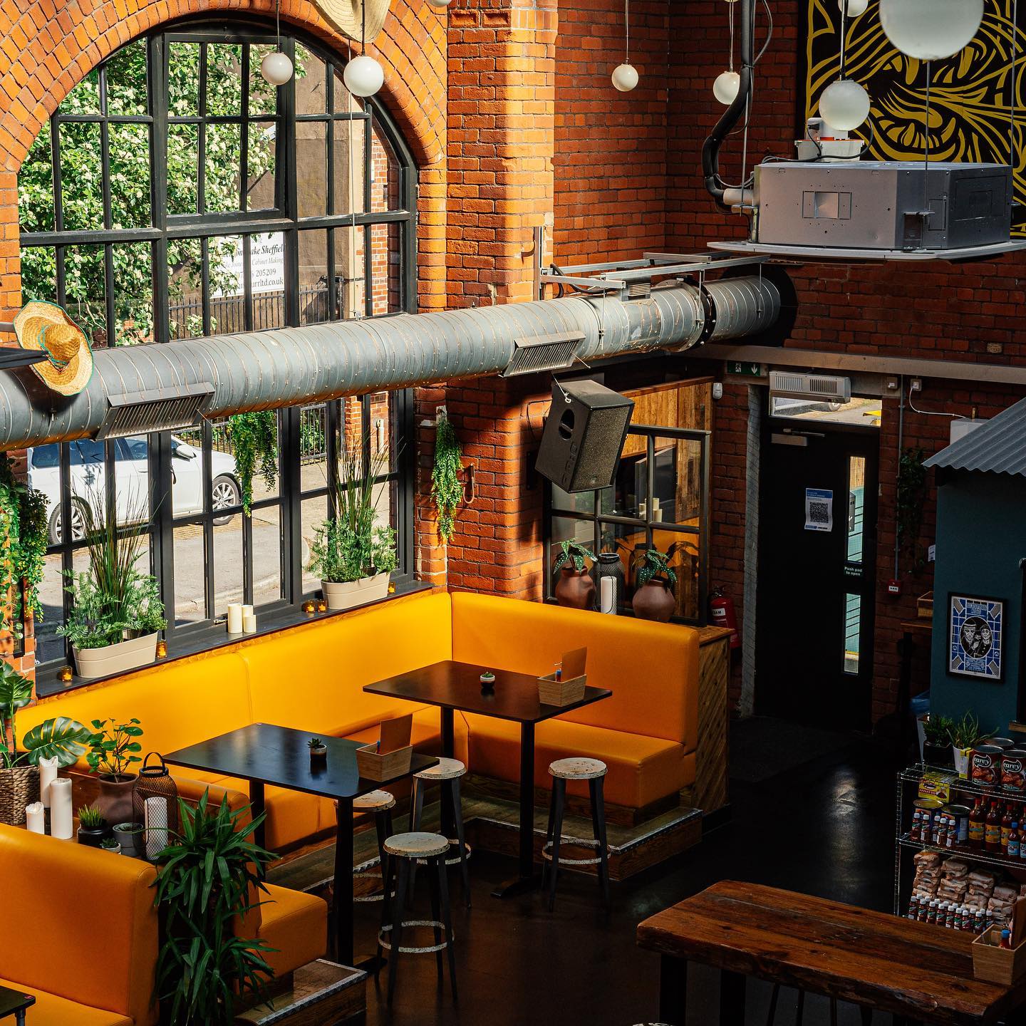 Industrial-style café interior with exposed red brick walls, large black-framed windows, and a prominent metal air duct running across the ceiling. Bright yellow booth seating surrounds dark wooden tables with stools. Green potted plants decorate the space, adding a natural touch. Overhead, round pendant lights hang from the ceiling, and a patterned artwork is visible on the upper wall. A door and a small retail section with snacks are in the background.