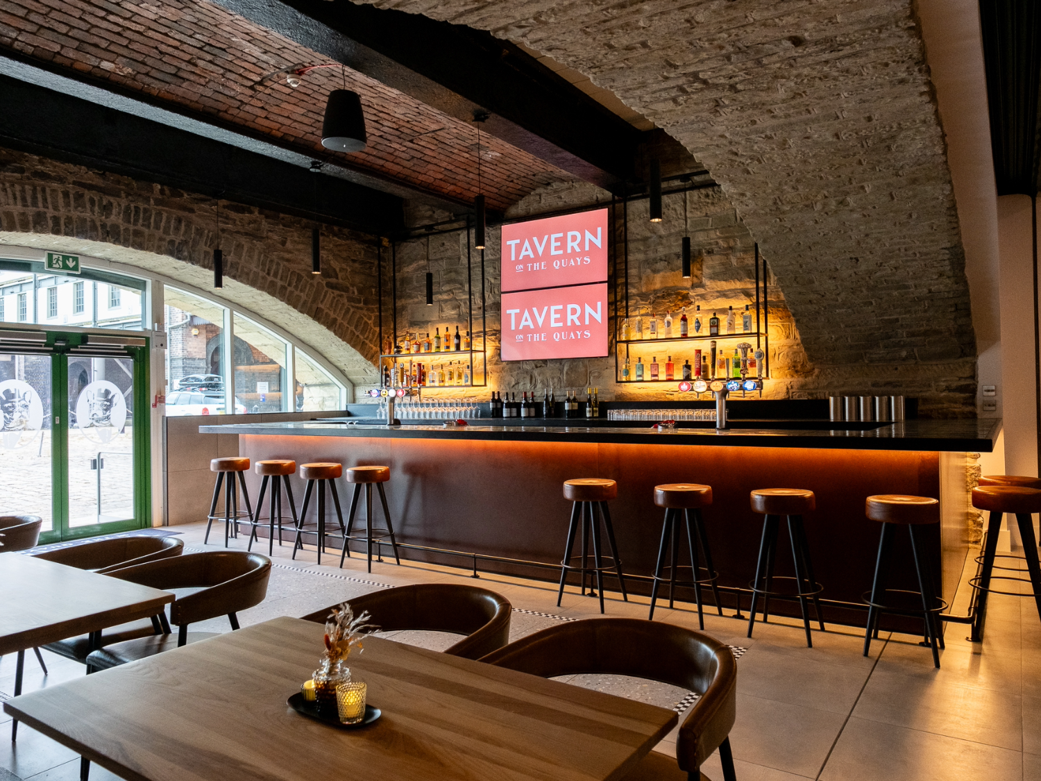 The bar at Tavern on The Quays, with the vaulted stone ceiling plus bar stools lined up along the bar and tables and chairs in the foreground.
