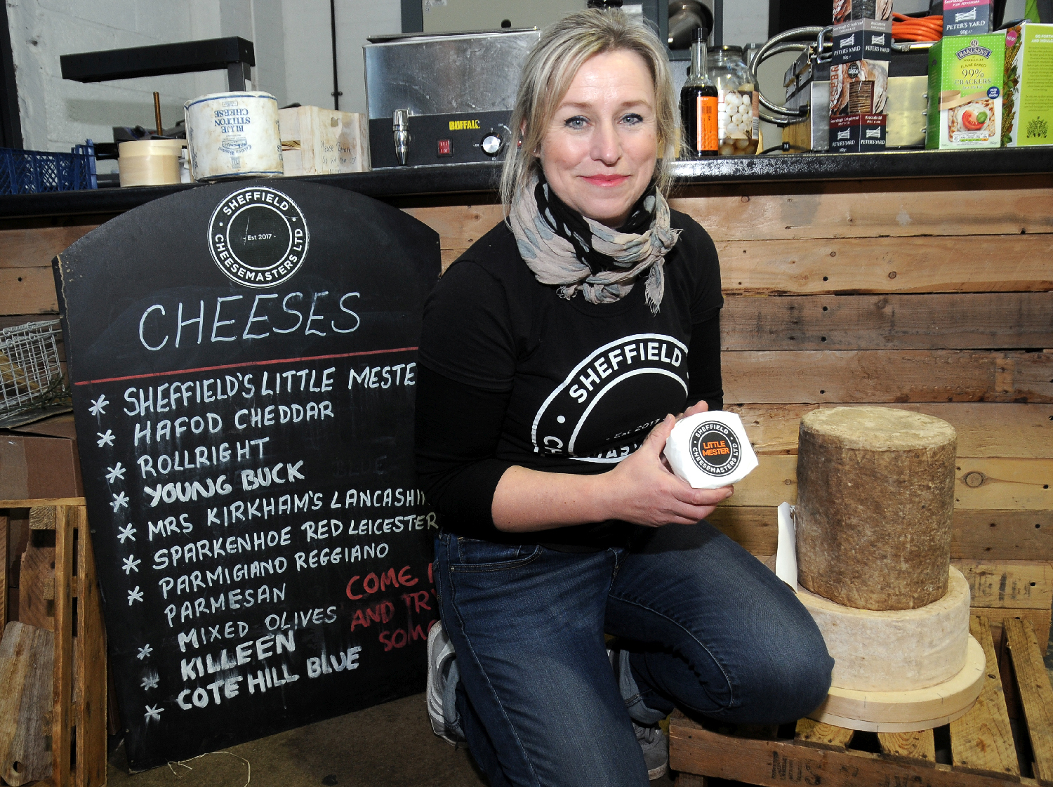 A business owner kneels on the floor of her Sheffield cheese shop, next to a blackboard, listing types of cheese and holding a block of cheese.