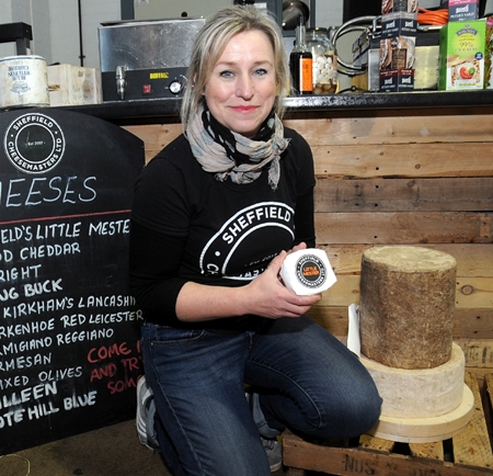 A business owner kneels on the floor of her Sheffield cheese shop, next to a blackboard, listing types of cheese and holding a block of cheese.