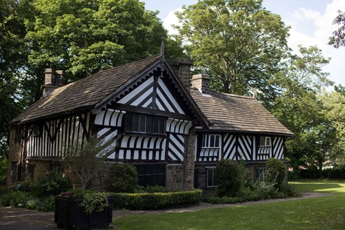 A historic black-and-white timber-framed building with a steep tiled roof sits in a green park surrounded by tall trees. The structure features decorative diagonal beams, small leaded windows, and stone walls at the base. A manicured lawn and shrubs frame the front, with a paved path leading toward the building under a bright, partly cloudy sky.