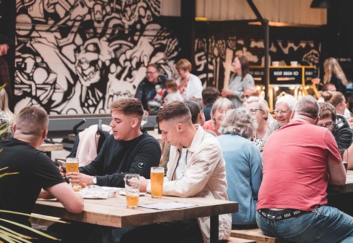 Crowds of people sat on benches drinking beer indoors, with a black and white graffiti wall in the background
