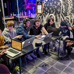Eight people, all wearing headphones, are sitting round a large table working on four laptops.