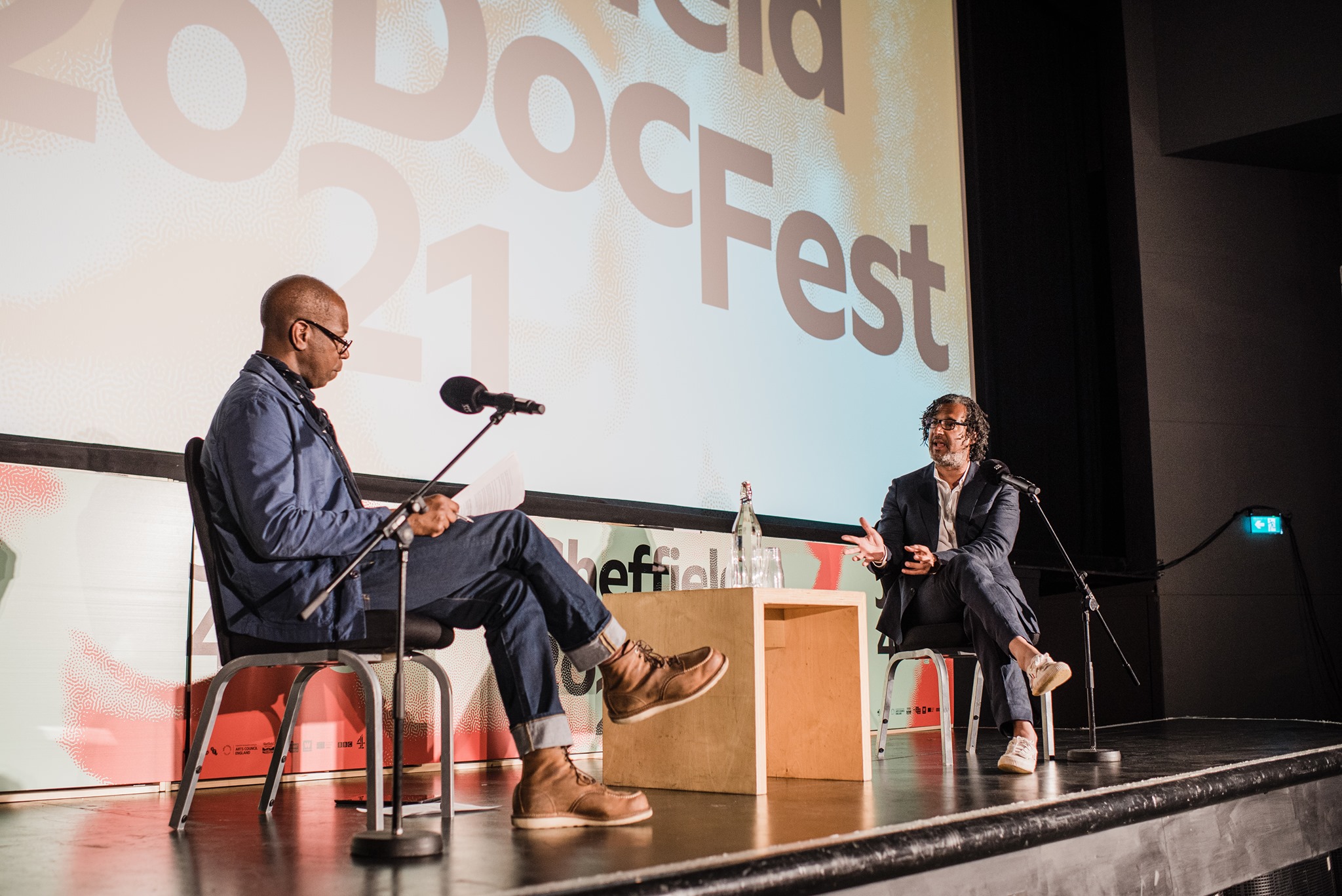 Two people seated on stage during a discussion at Sheffield DocFest, with microphones on stands and a small wooden table between them. A large screen behind displays the event name and year.