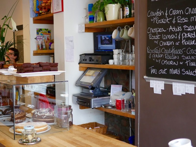 A counter in a cafe, with a glass case filled with cakes. Behind the counter is a menu written on a large blackboard.