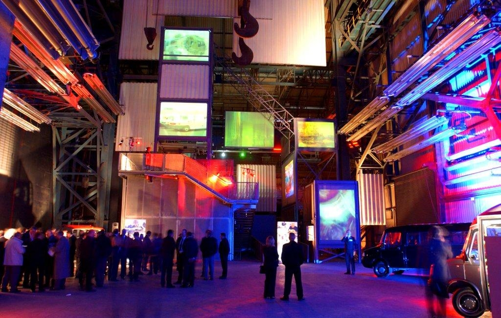 The Face of Steel - Interior of an industrial-style venue with high ceilings and exposed metal beams, illuminated by colorful red, blue, and purple lights. Several large screens display visuals, and groups of people are gathered around the space. A staircase and suspended structures add to the futuristic atmosphere.