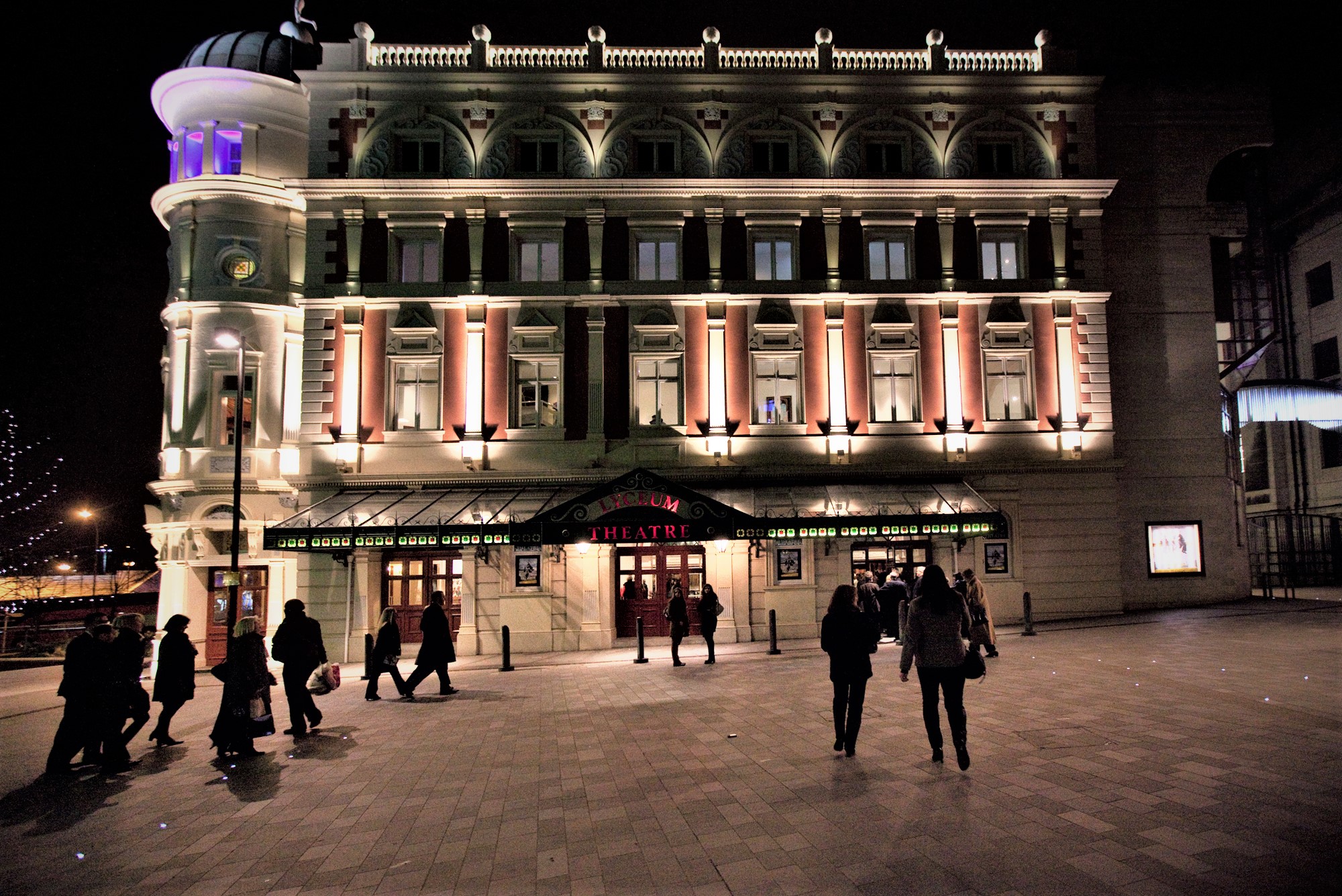People walking through Tudor Square towards the Lyceum Theatre, at night.