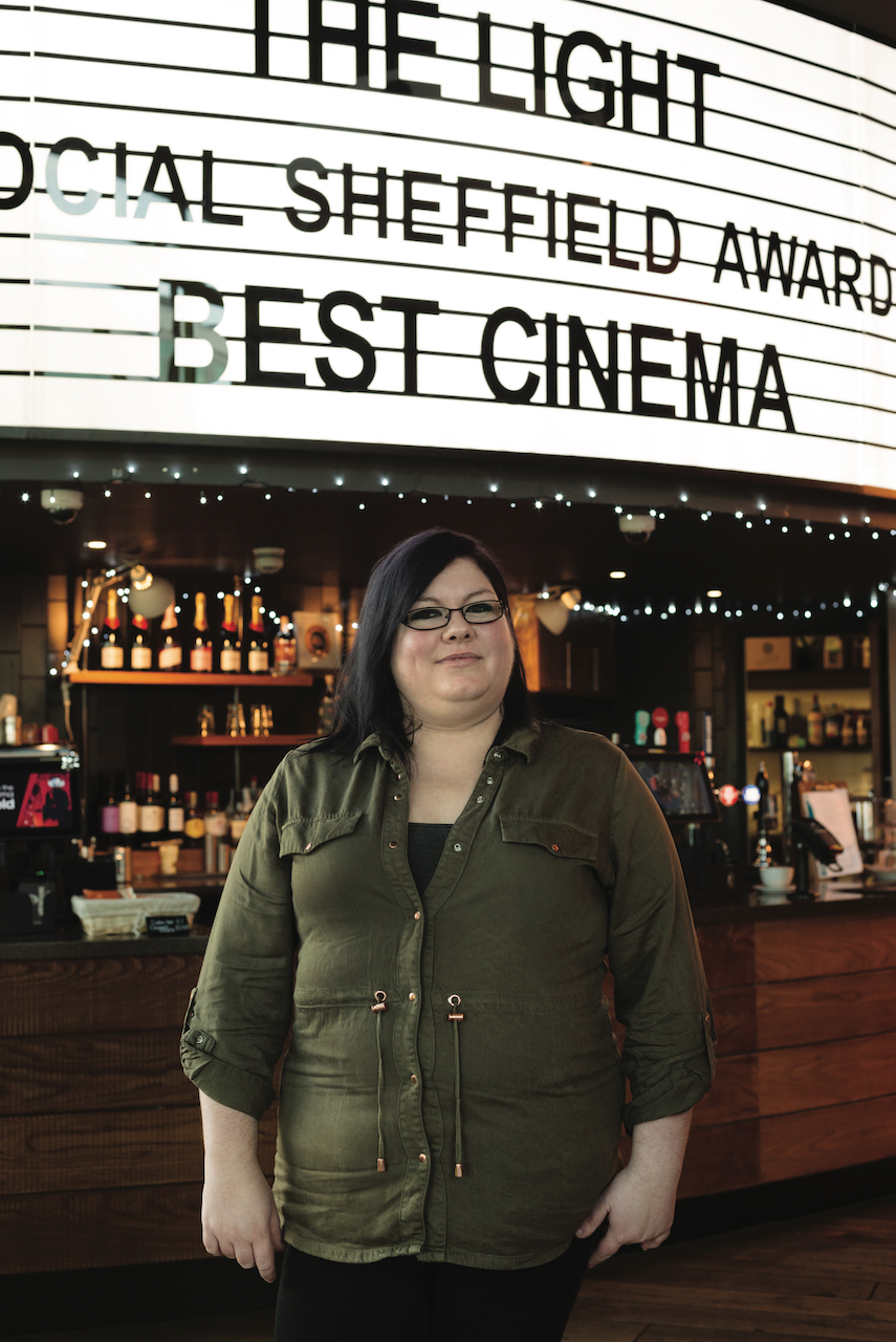 Person standing in front of a bar counter inside a cinema, with a large illuminated sign overhead reading ‘THE LIGHT SOCIAL SHEFFIELD AWARD BEST CINEMA.’ Shelves behind the counter display bottles and glasses, and string lights add decoration.