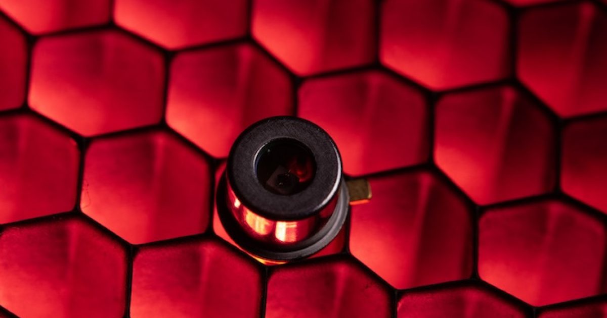 Close-up of a small cylindrical electronic component mounted on a surface with a red honeycomb pattern. The component has a metallic finish and a central lens-like opening, reflecting the red light from the background.