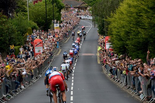 Cyclists racing along a closed road lined with metal barriers and large crowds of spectators on both sides, during a professional cycling event in an urban area.