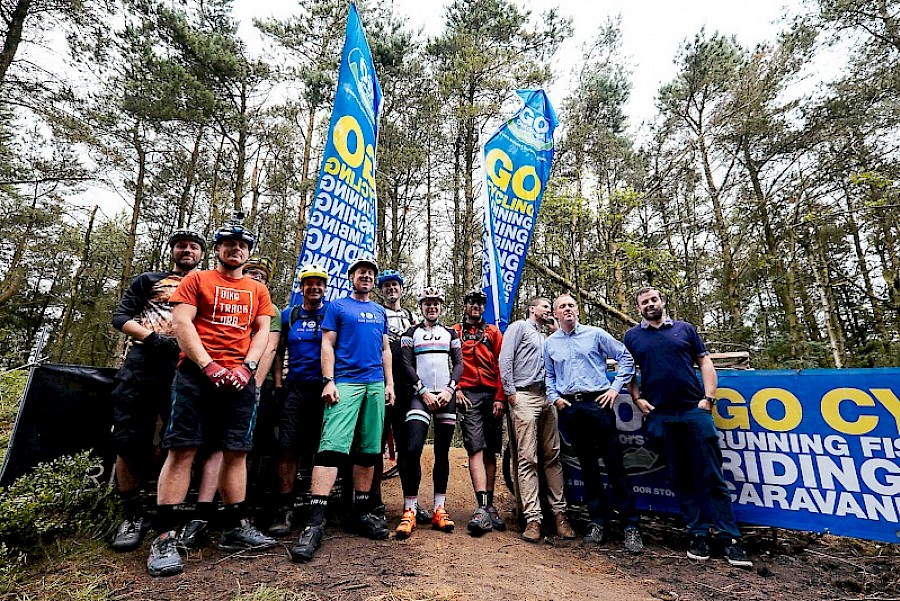 A group of people stands on a forest trail in front of two tall blue flags and a large banner that reads “GO CY” with words like “RUNNING,” “RIDING,” and “CARAVAN” visible. The individuals are dressed in casual and cycling gear, with some wearing helmets and gloves. The background shows tall trees and greenery, suggesting an outdoor cycling or adventure event.