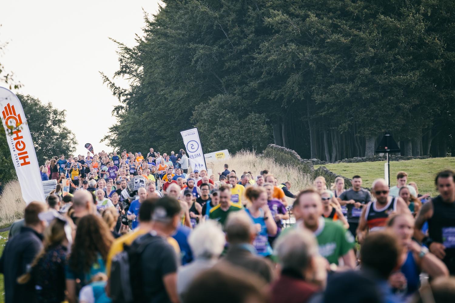 A large crowd is watching and cheering on runners running on a road. In the background is a large wood.
