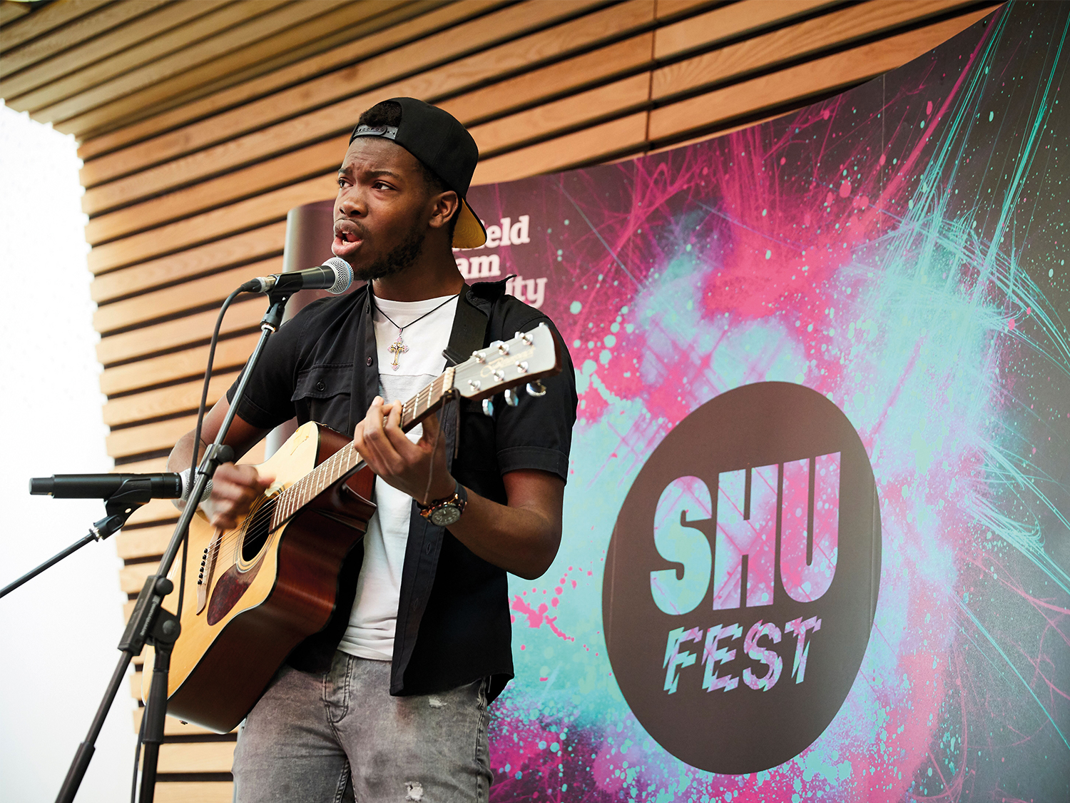 A person is performing on stage, playing an acoustic guitar and singing into a microphone. The backdrop features a colorful design with the text “SHU FEST” and “Sheffield Hallam University.” The setting appears to be an indoor event space with wooden panelling and stage lighting.