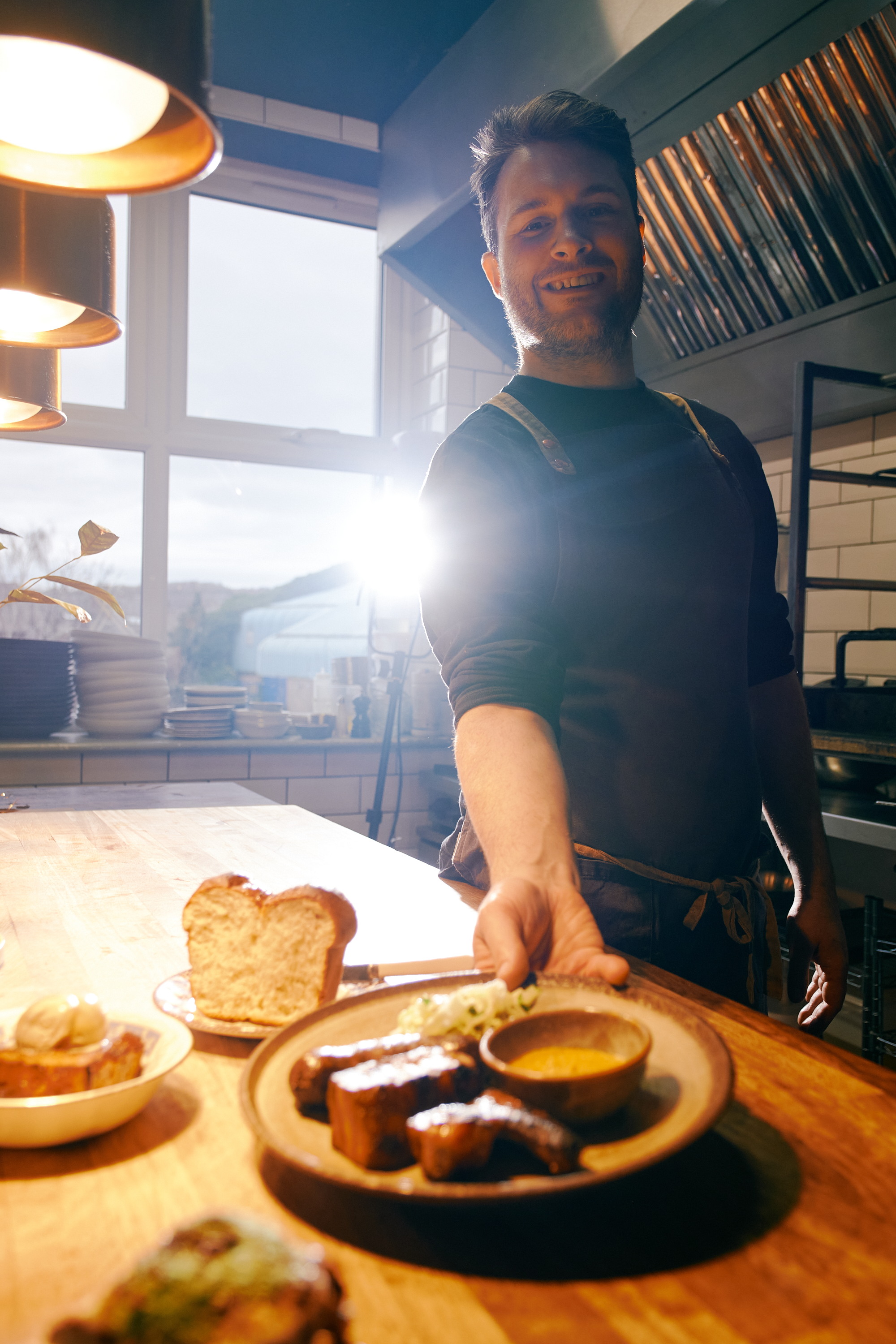 A smiling man, in an apron, is placing a plate of food down on a table.