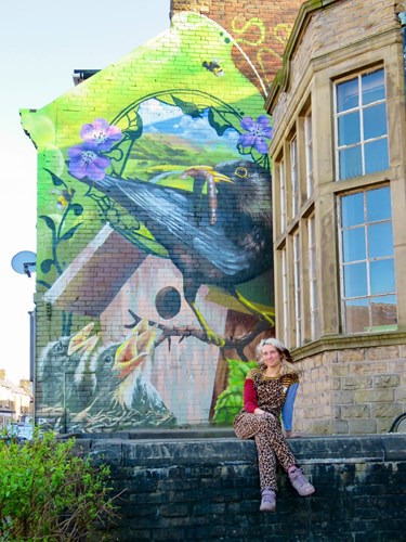 Large mural painted on the side of a brick building depicting a blackbird holding a worm in its beak, perched near a wooden birdhouse with three chicks inside. The mural includes green foliage and purple flowers against a bright background. In the foreground, a person wearing a patterned outfit and pink shoes is sitting on a stone ledge next to the building’s bay window.