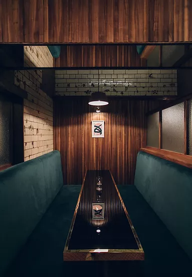 A cozy booth in a dimly lit bar or restaurant with teal upholstered seating on both sides of a long, glossy black table. The walls feature a mix of vertical wooden panels and white subway tiles with dark grout. A single pendant light hangs above the table, casting a warm glow. On the back wall, there is a framed minimalist artwork. The overall design combines modern and rustic elements, creating an intimate atmosphere.