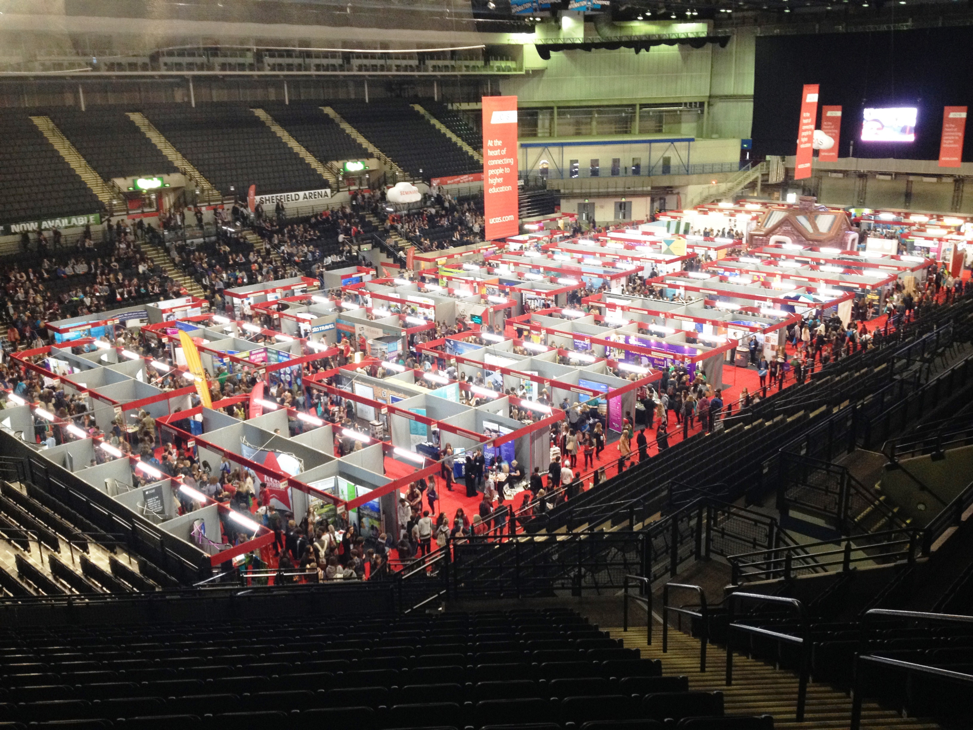 Large indoor exhibition at Sheffield Arena with rows of booths arranged on a red carpeted floor, surrounded by crowds of visitors. Elevated seating areas are visible in the background.