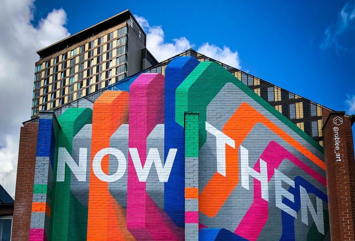 A vibrant mural on the side of a building displays the words 'NOW THEN' in large white letters against a colorful background of overlapping geometric shapes in blue, green, orange, pink, and purple. A tall building with many windows stands in the background under a partly cloudy sky. The artist's Instagram handle '@roblee.art' is visible on the right.