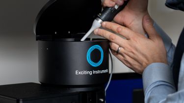 Close-up of hands using a pipette to place a sample into a black cylindrical scientific instrument labeled “Exciting Instruments” with a blue circular logo. The instrument lid is open, and part of a computer monitor is visible in the background.