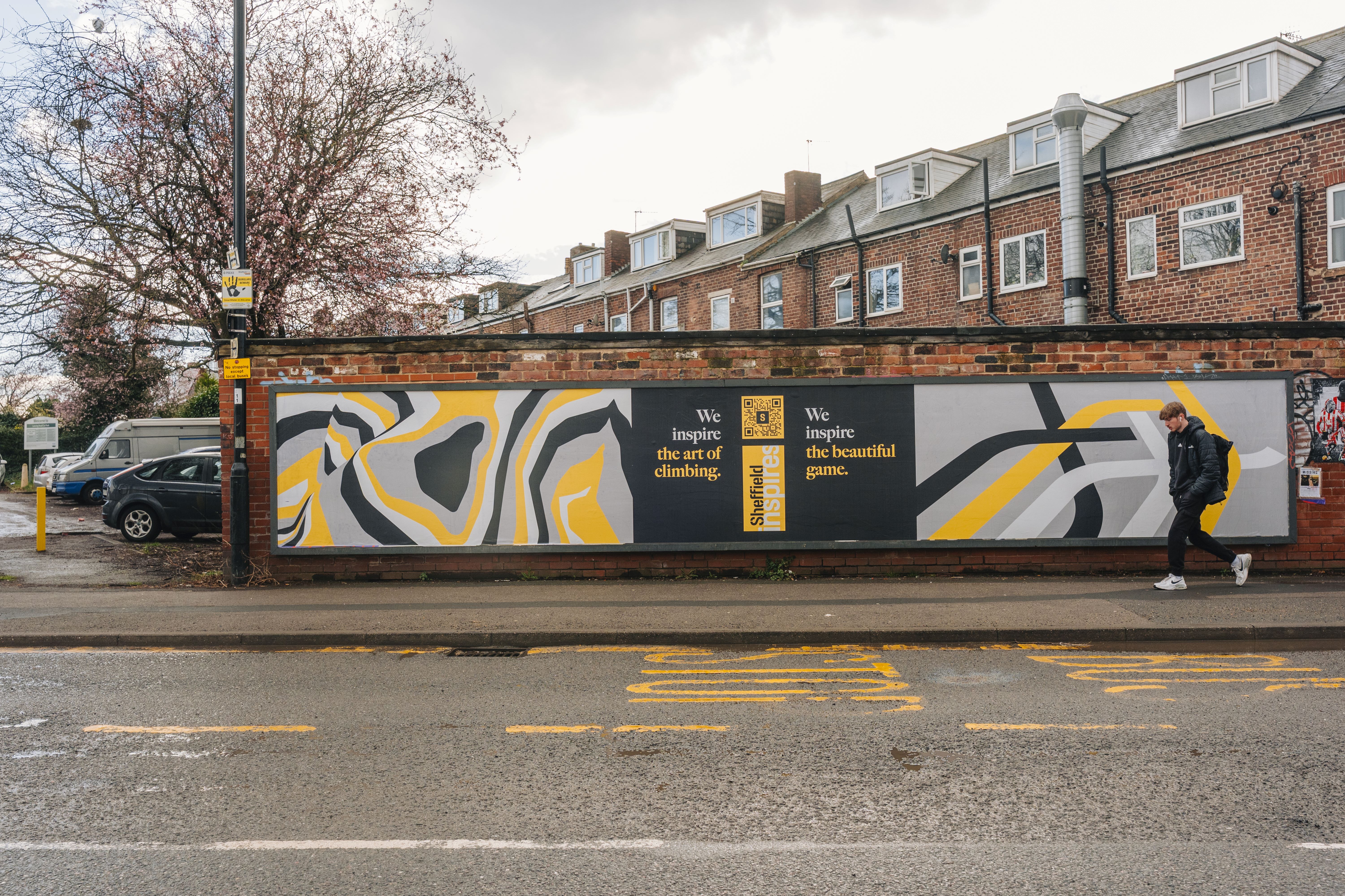A street mural painted on a brick wall featuring abstract patterns in black, white, gray, and yellow. The mural includes two text sections: “We inspire the art of climbing” and “We inspire the beautiful game.” A person wearing a hooded jacket and carrying a backpack walks past the mural on the sidewalk. Behind the wall are rows of red-brick houses with white-framed windows and sloped roofs. The street in front has faded yellow road markings and appears wet, suggesting recent rain.
