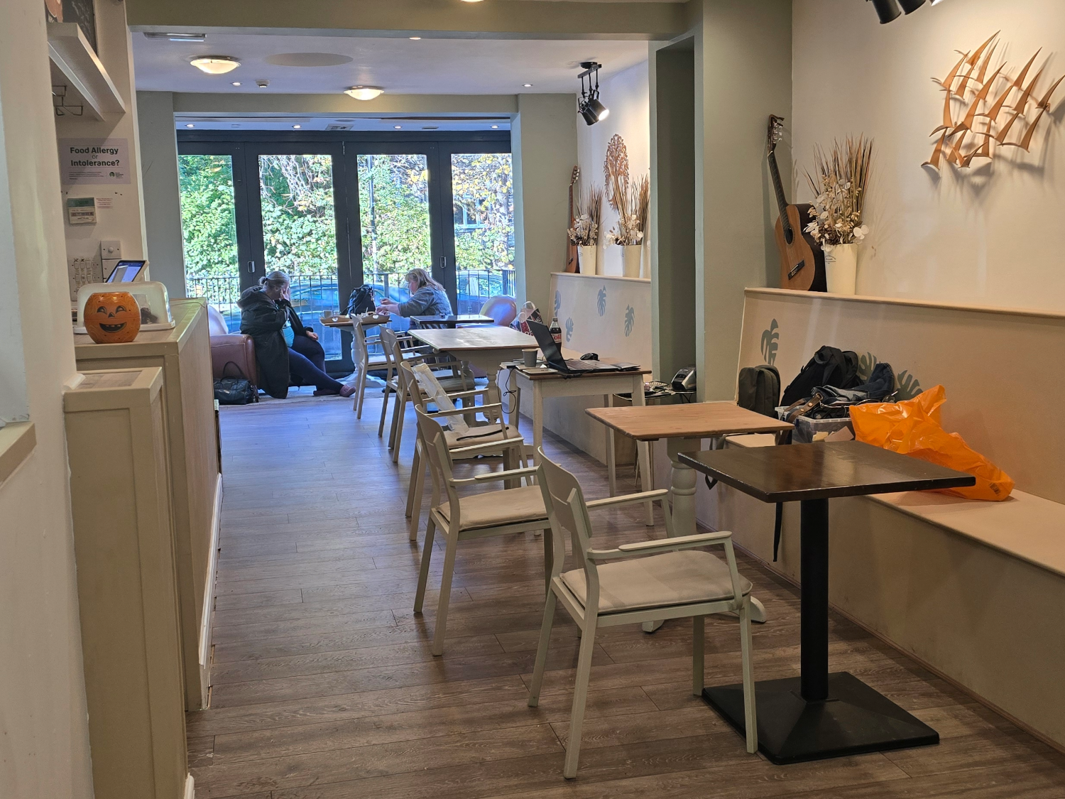 Interior of a cozy café with wooden flooring and light-colored walls decorated with dried floral arrangements and a mounted guitar. Several tables and chairs are arranged along the narrow space, with a bench running along the right wall. A small pumpkin decoration sits on a shelf to the left. Large glass doors at the back open to a view of trees outside, and a few people are seated near the windows.