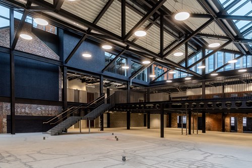 Interior of a large industrial-style building with exposed steel beams, high ceilings, and modern lighting. The space features a mezzanine level with black railings, a staircase leading up, and a mix of brick and metal walls. The floor is unfinished concrete, and large windows allow natural light into the open area.