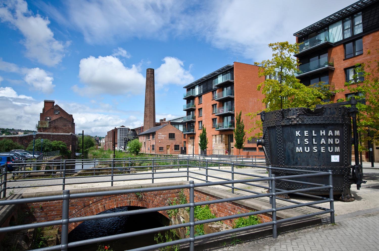 An outdoor view of Kelham Island Museum in Sheffield, featuring a large black industrial structure with the museum name in white letters. The scene includes modern red-brick apartment buildings, an old factory chimney in the background, and a canal running under a brick arch bridge with metal railings. The sky is bright blue with scattered clouds, creating a vibrant urban-industrial setting.
