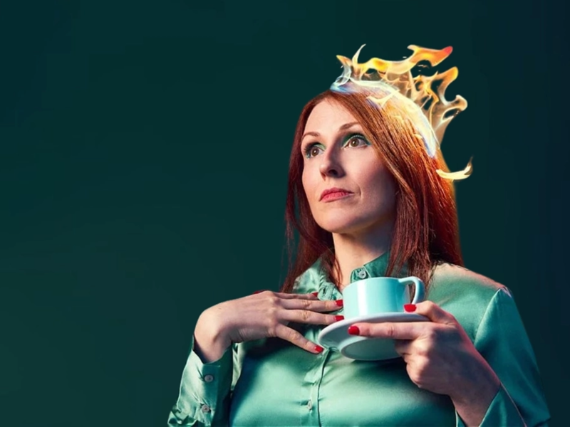 A woman, looking distracted, in a green dress holding a cup of tea. There are flames on her hair.