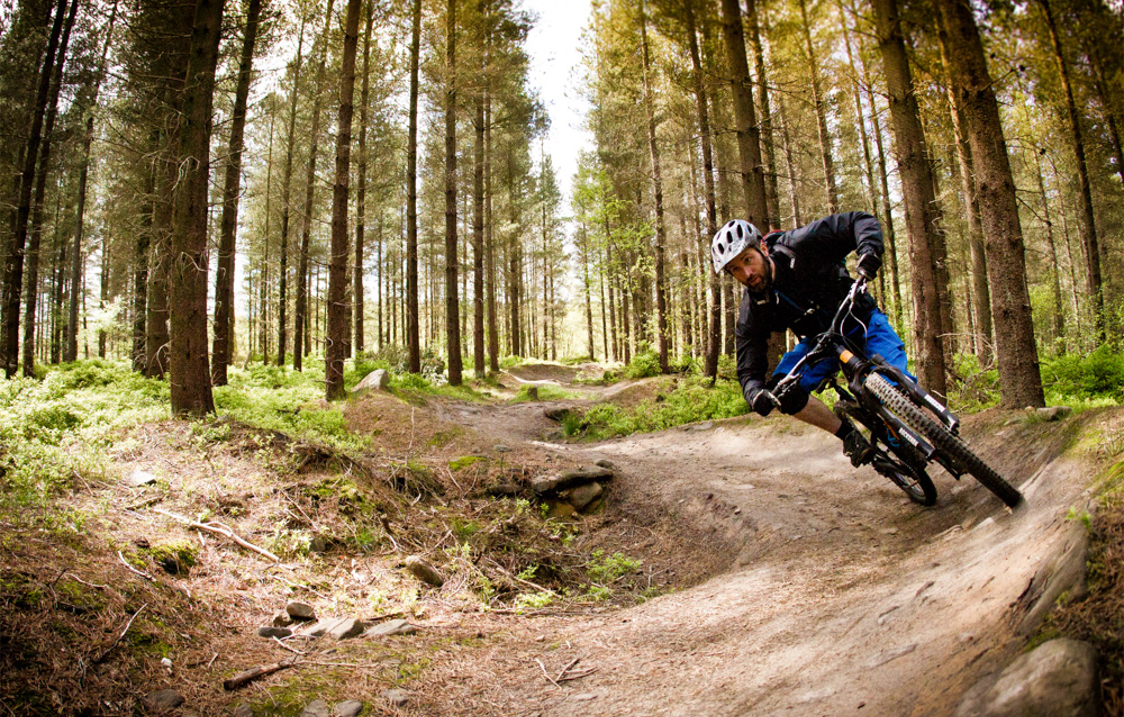 A man on a mountain bike cycles round a banked curve on a dirt track in a wooded area.