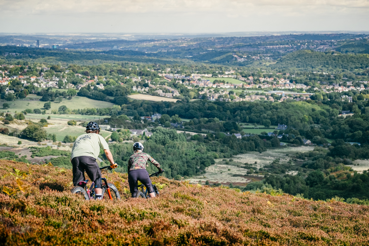 Two people are mountain biking along a dirt track, flanked by heather, on a hillside above Sheffield.
