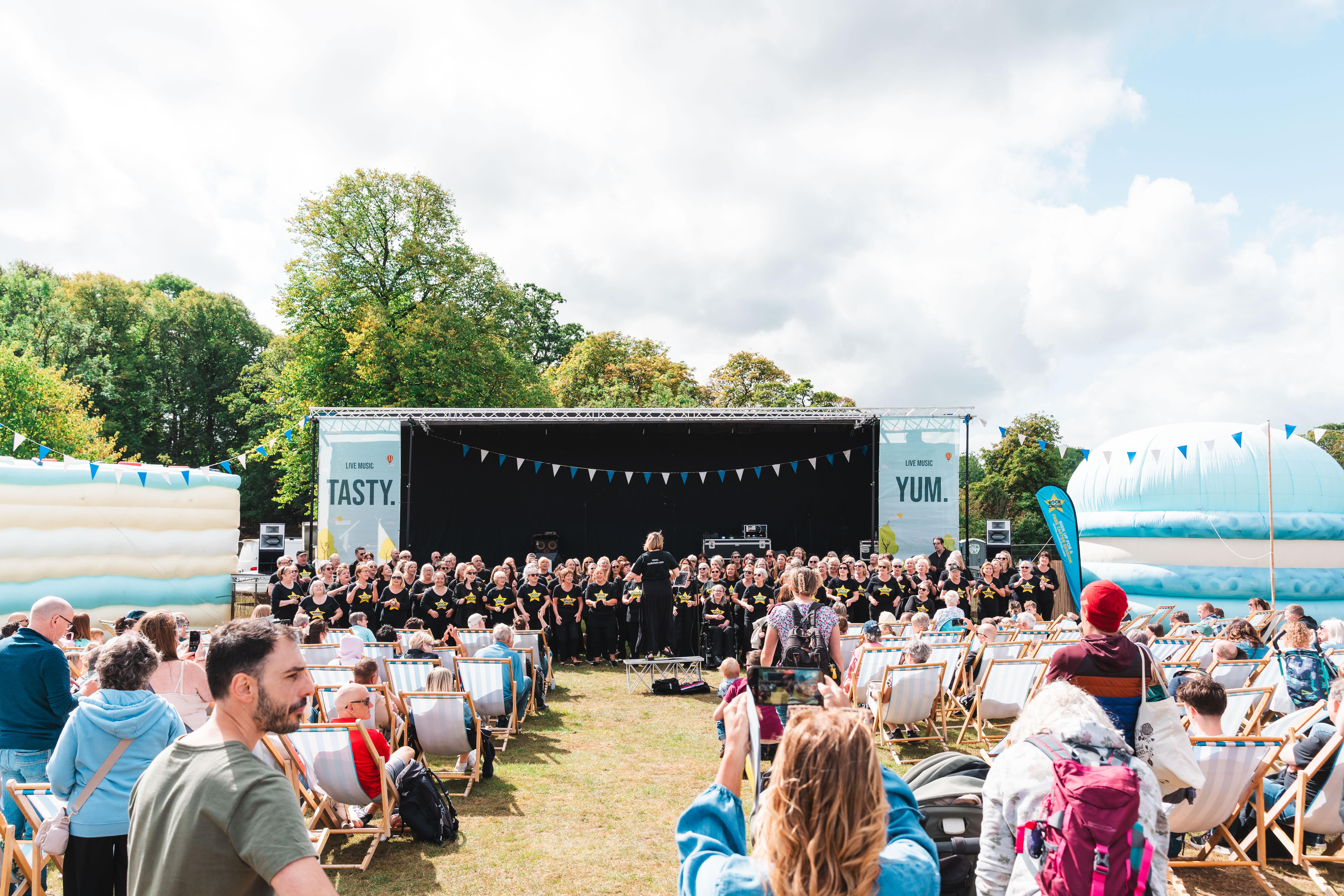 Outdoor event with a large stage featuring a choir performance, rows of deck chairs filled with spectators, and colourful bunting overhead. Inflatable structures and trees surround the area under a partly cloudy sky.